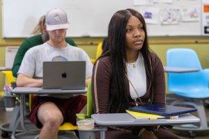 A high school girl sitting at a desk in a classroom with other students sitting at desks behind her.