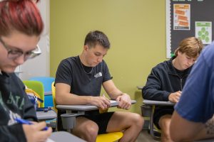 Three high school students sitting at desks in a classroom looking down at papers.