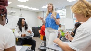 A teacher standing in front of a small group of three students who are sitting at desks in a classroom.