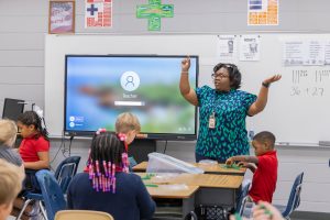 A teacher standing in front of elementary students.in a classroom with arms raised and students conducting activity at desks.