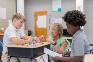 Two student grouped together at a desk with a teacher crouched down at the desk to help them with their class assignment.