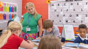 A teacher standing in a classroom helping a group of four students with an assignment at their desks.