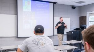 A male professor presents at the front of a classroom near a podium and projection screen, with two students visible in the foreground.