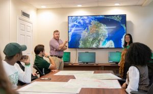 A student and professor, one standing on each side of a large TV screen mounted on the wall with a map on it, in front of students sitting at a conference table that has physical maps on it.