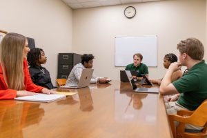 A group of six students sitting around a conference table with laptops and notebooks on the table as they engage in a discussion.