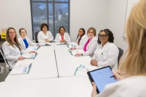 A group of MSN students in lab coats sit around a conference table, facing a person with a tablet who appears to be leading or presenting during a meeting.