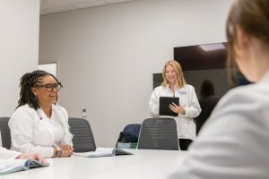 Three women in lab coats in a discussion during a meeting, two seated and the other standing with a tablet.