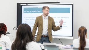 A male professor stands in front of a screen giving a presentation to a group of people in lab coats.