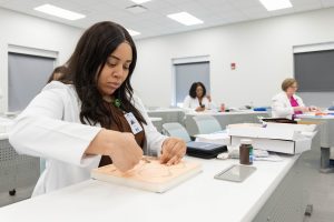 Woman in a lab coat practicing suturing on training pad in a classroom, with others working in the background.