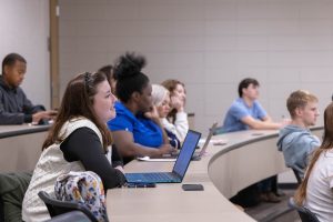 Students seated in a classroom, attentively listening to a lecture with laptops and open notebooks in front of them.