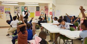 Elementary-aged students in a brightly decorated classroom raising their hands while a college student speaks at the front of the room. Some students are actively writing or listening.