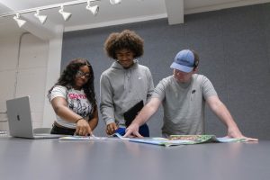 Students collaborating on a creative project, pointing to pages of printed work laid out on a table with a laptop in the background.