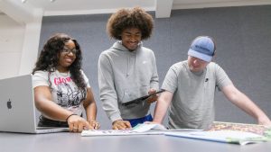 Students collaborating on a creative project, pointing to pages of printed work laid out on a table with a laptop in the background.