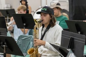 University band student playing saxophone in rehearsal, surrounded by classmates performing on trumpets and percussion.