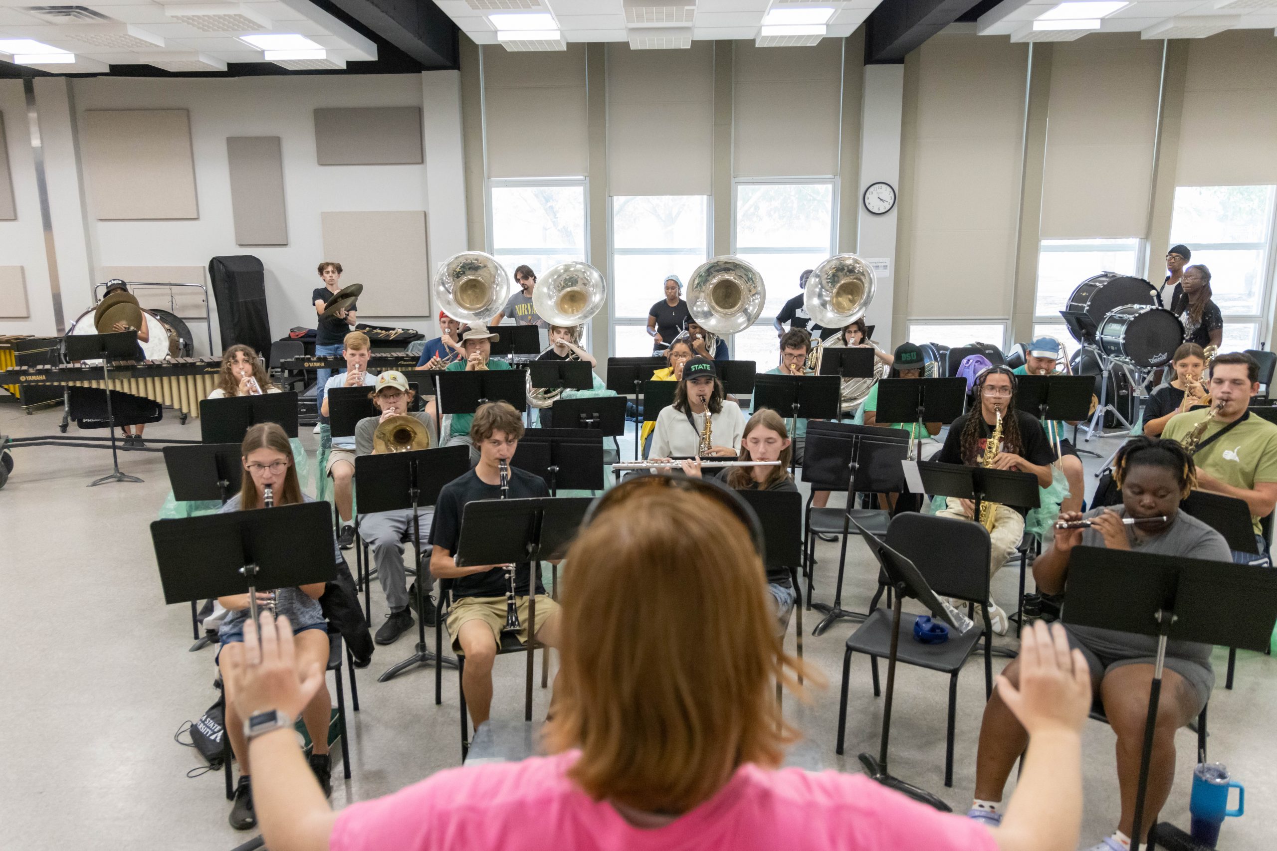 University band students rehearsing under the direction of a conductor, with woodwinds, brass, and percussion instruments filling the classroom.