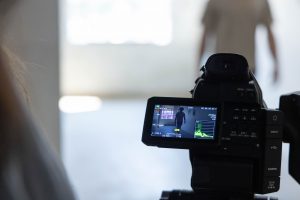 Film production student recording a class project with a professional video camera, capturing another student on screen.