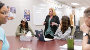 Instructor leading a discussion with students seated around a table in a classroom.