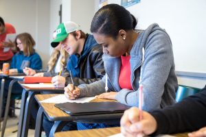 Students sitting at desks in a classroom, writing on paper. A woman in a gray hoodie and red shirt is focused on her work beside a man in a green and white cap.