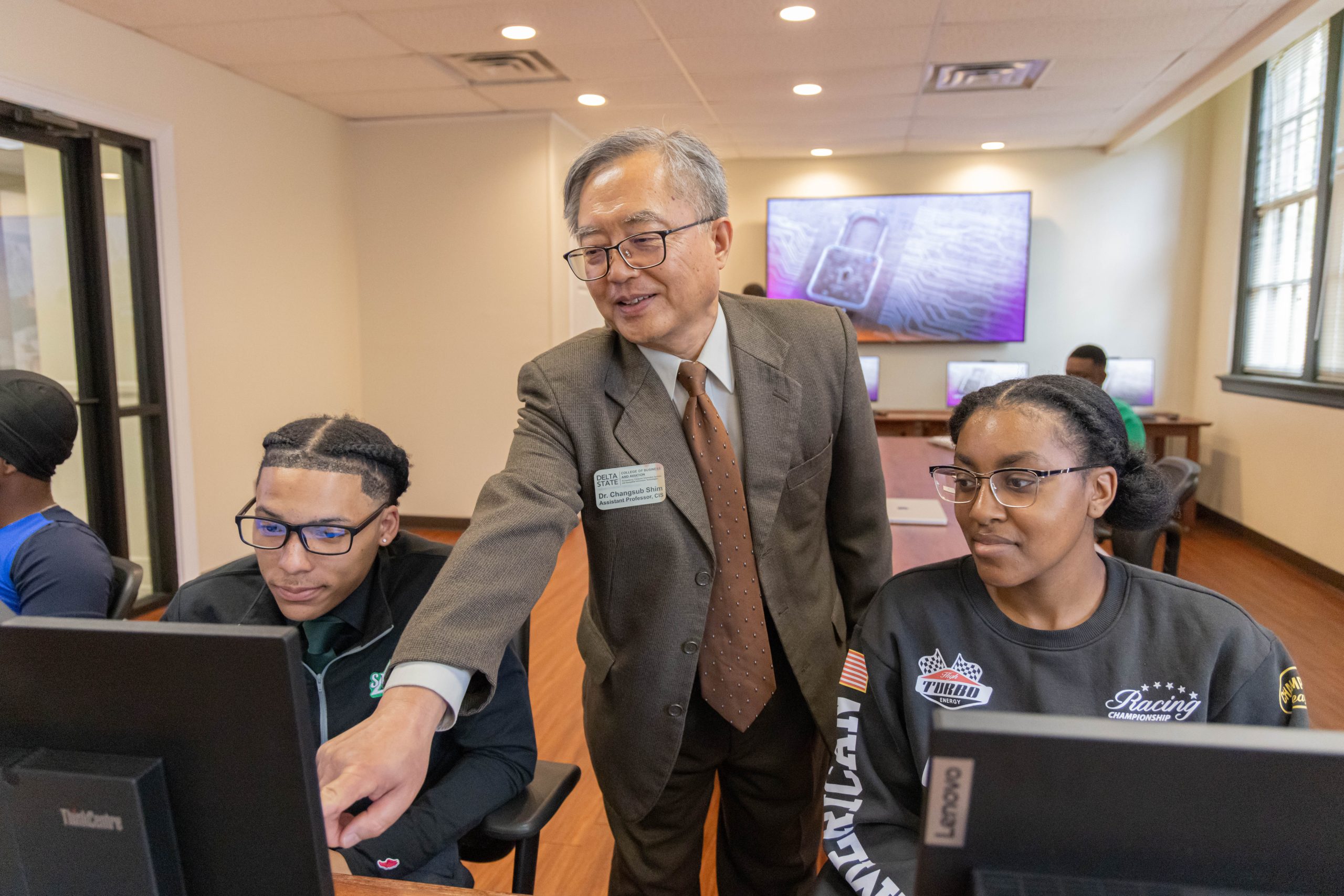 Instructor assisting two students at desktop computers in a classroom, pointing at a screen while they work.