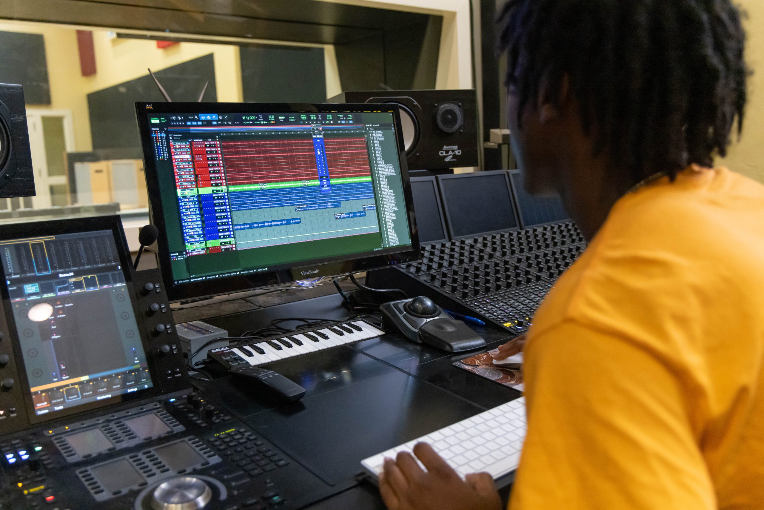 Student working at a music production workstation, editing audio tracks on a computer monitor with a mixing console, MIDI keyboard, and studio equipment in a recording studio.