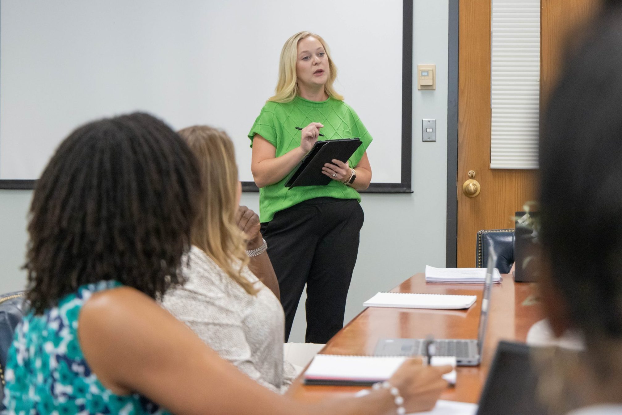 A woman in a green shirt stands at the front of a classroom holding a tablet and speaking, while several students sit at a table facing her with notebooks and materials.