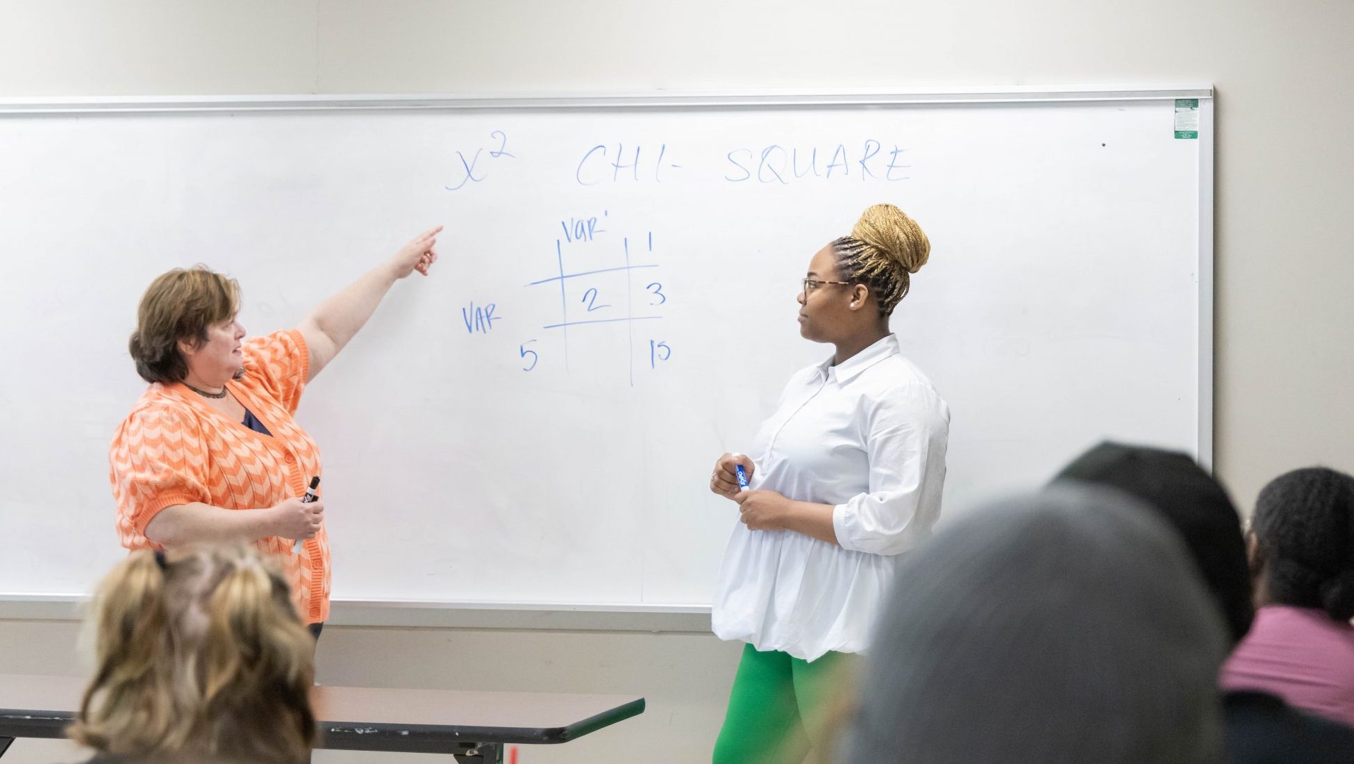 Professor and Graduate Assistant at a whiteboard teaching a classroom of students, with one pointing to a chi-square equation.
