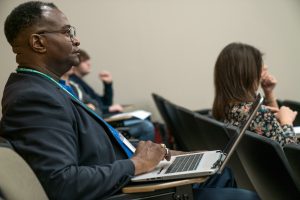 A man in a blazer attentively listens during a lecture or presentation, seated with a laptop on his lap in a lecture hall with other attendees nearby.