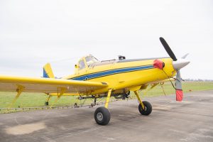 A yellow crop-dusting airplane parked on the tarmac with spray booms attached beneath the wings.