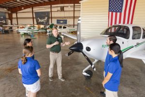 Instructor leading a lesson with aviation students next to a training aircraft inside a hangar.
