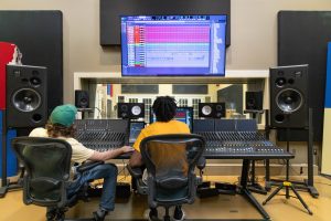 Two students operating a digital mixing console in a professional recording studio with monitors and speakers.