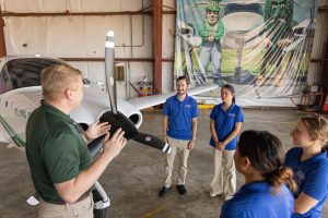 Delta State University aviation instructor speaking with students beside a training aircraft inside a hangar.