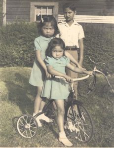 Brother and two sisters posing near tricycle. Color.