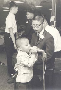 Little boy at microphone, standing next to older boy. B&W.