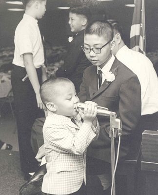 Little boy at microphone, standing next to older boy. B&W.