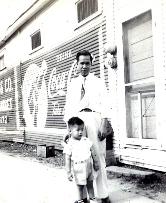 Man poses with child, outside of storefront. B&W.