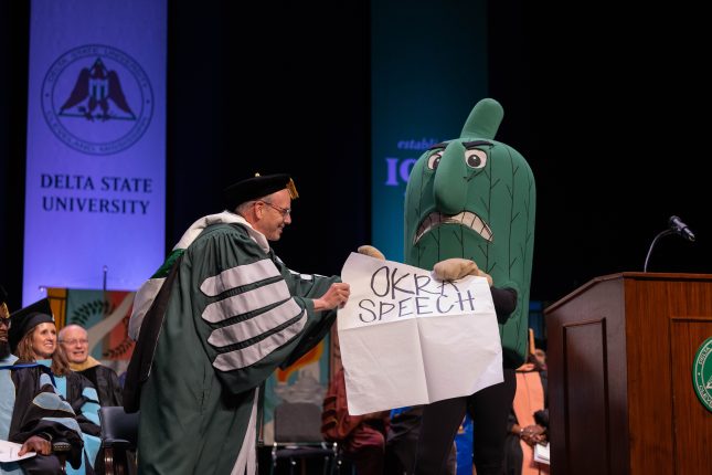The Fighting Okra mascot with a large piece of paper reading "Okra Speech" being assisted by DSU President Dan Ennis