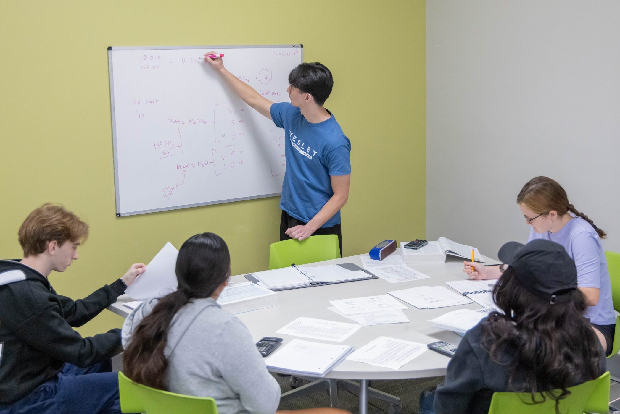 Student leading a small group study session at a whiteboard while others take notes around a table.