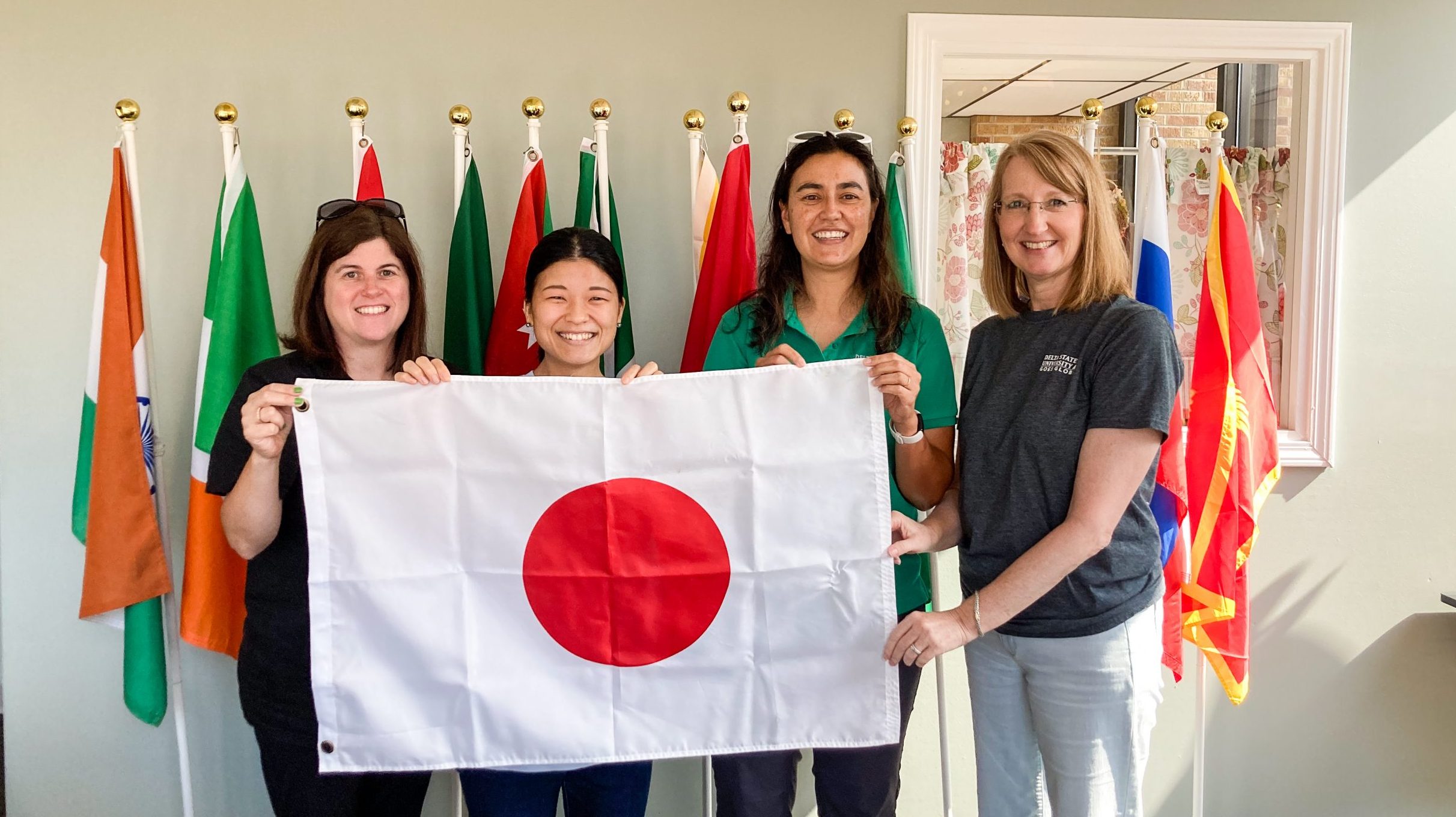 Four women smiling and holding the Japanese flag in front of a display of international flags.
