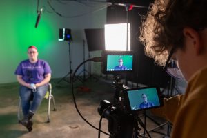 Student sitting in a chair with video equipment and lighting placed around her, while another student operates a camera on a tripod with visible displays.