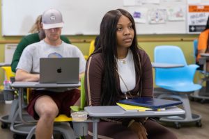 A high school girl sitting at a desk in a classroom with other students sitting at desks behind her.