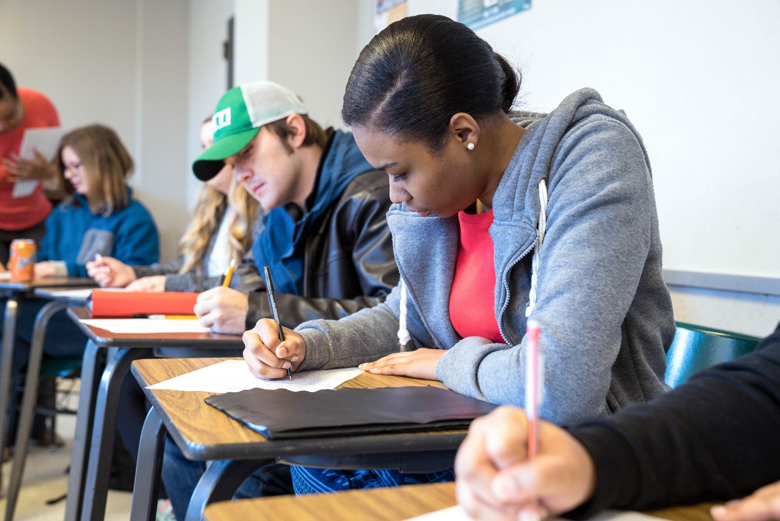 Students sitting at desks in a classroom, writing on paper. A woman in a gray hoodie and red shirt is focused on her work beside a man in a green and white cap.