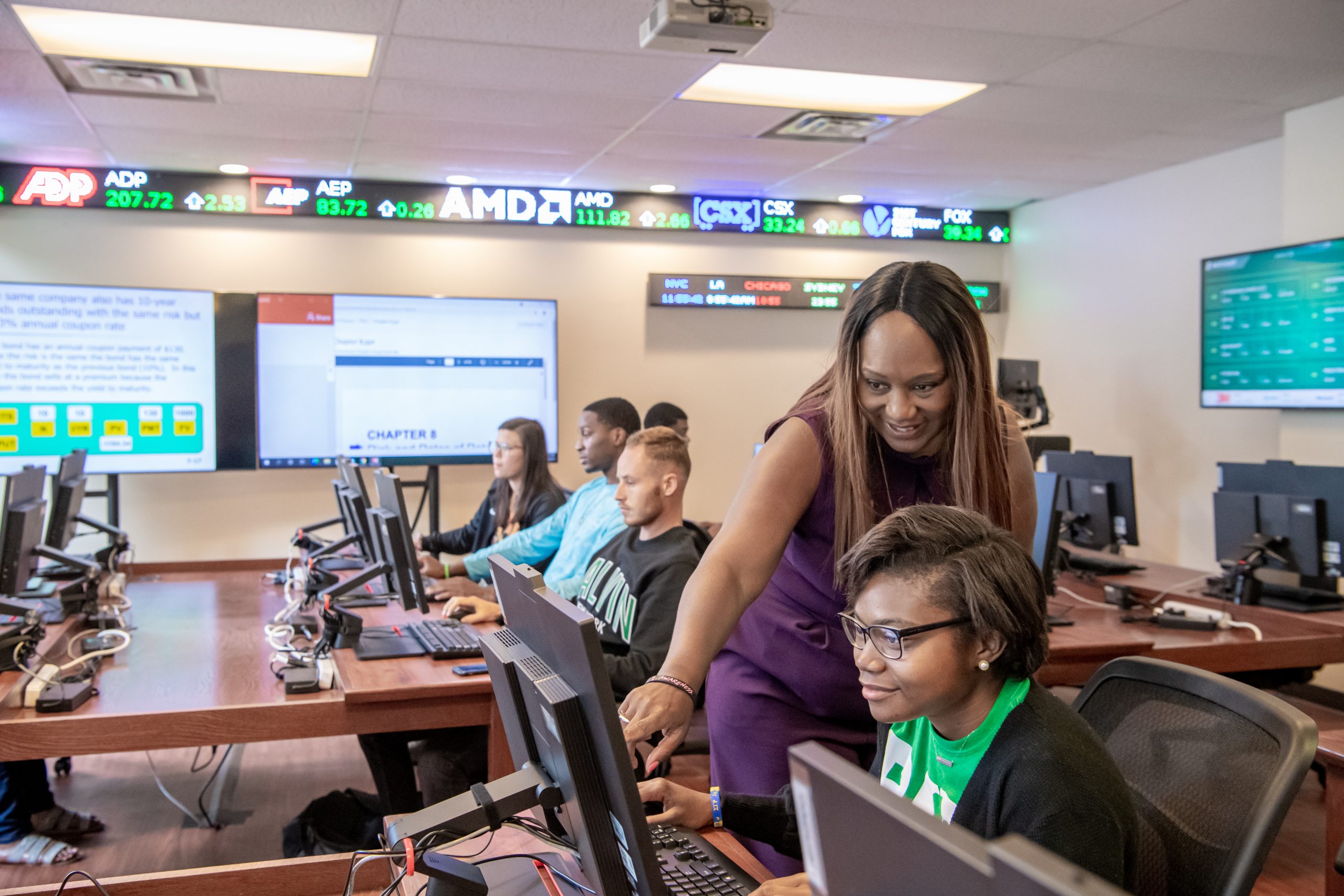Business instructor helping a student at a computer while classmates work in finance lab classroom.